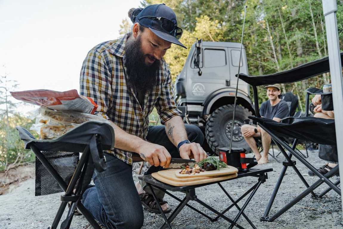 Guest slicing vegetables at a camp table beside a Mercedes-Benz expedition truck