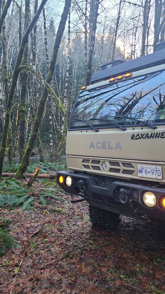 Acela expedition truck with its lights on in a dark forest at dusk