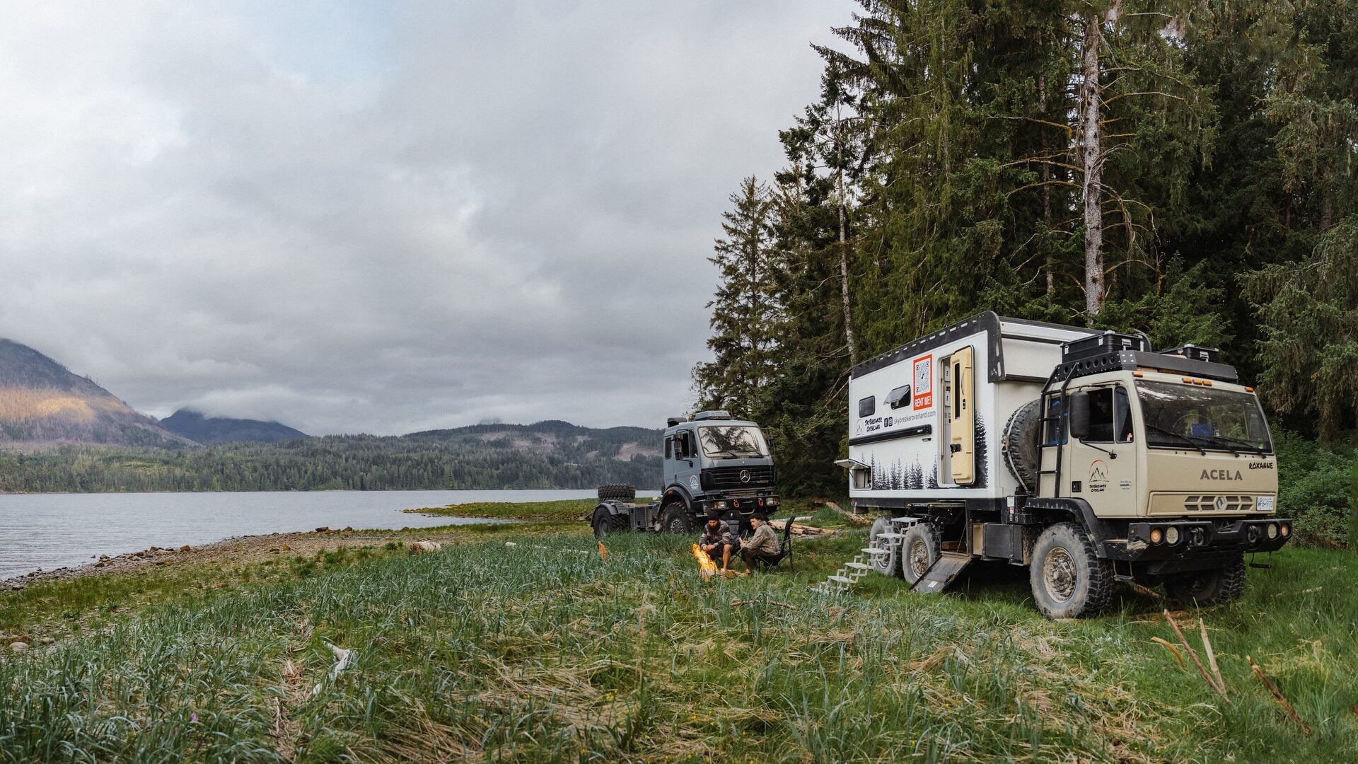 Roxanne camped on grass above a lake, entry steps down and a fire burning between the two trucks